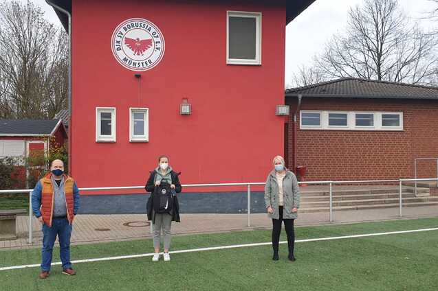 Drei Personen stehen vor einem roten Vereinsgeb&auml;ude, umgeben von einem Fu&szlig;ballfeld mit Kunstrasen.