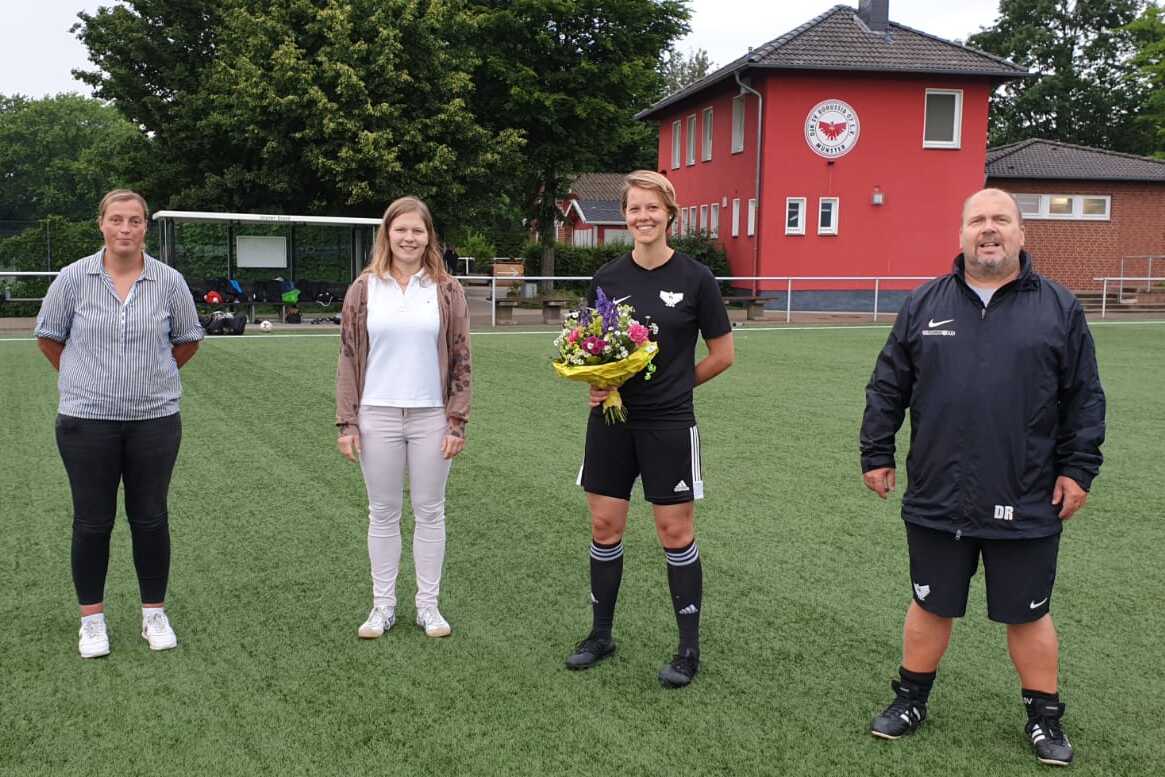 Vier Personen stehen auf einem Fu&szlig;ballfeld, eine Spielerin h&auml;lt einen Blumenstrau&szlig;, im Hintergrund ein rotes Vereinsgeb&auml;ude.