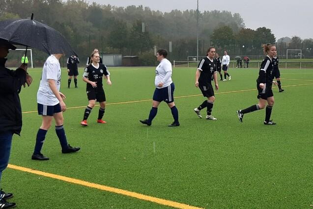 Fu&szlig;ballspiel bei Regen: Spielerinnen in wei&szlig;en und schwarzen Trikots laufen &uuml;ber den Kunstrasenplatz, Zuschauer mit Regenschirm.