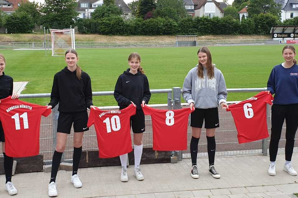 F&uuml;nf Sportlerinnen in Fu&szlig;balltrikots halten rote Shirts mit den Nummern 11, 10, 8 und 6 auf einem Fu&szlig;ballplatz.