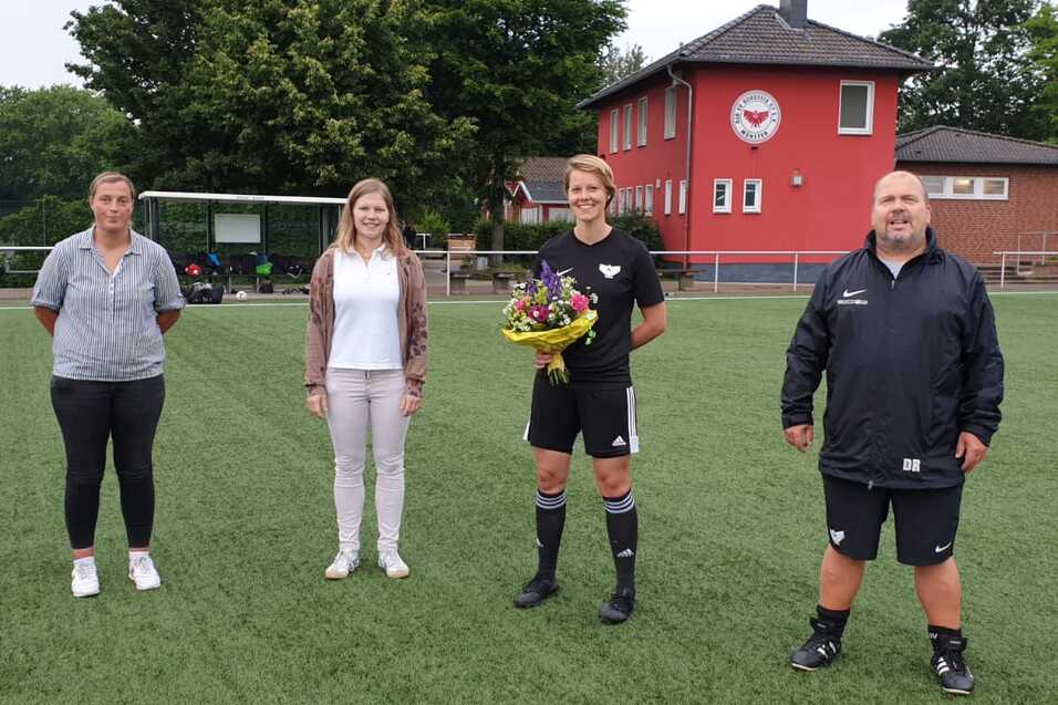 Vier Personen stehen auf einem Fu&szlig;ballfeld, eine Spielerin h&auml;lt einen Blumenstrau&szlig;, im Hintergrund ein rotes Vereinsgeb&auml;ude.