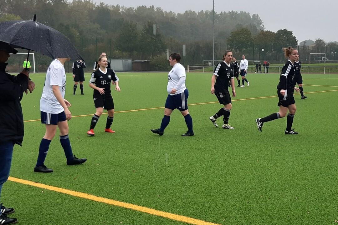 Fu&szlig;ballspiel bei Regen: Spielerinnen in wei&szlig;en und schwarzen Trikots laufen &uuml;ber den Kunstrasenplatz, Zuschauer mit Regenschirm.