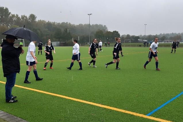 Fu&szlig;ballspiel bei Regen: Spielerinnen in wei&szlig;en und schwarzen Trikots laufen &uuml;ber den Kunstrasenplatz, Zuschauer mit Regenschirm.