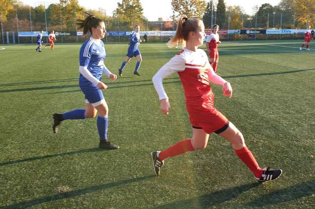 Eine Fu&szlig;ballspielerin in roter Uniform f&uuml;hrt mit einem Ball einen Kick auf einem Kunstrasenplatz aus, Spielerinnen im Hintergrund.