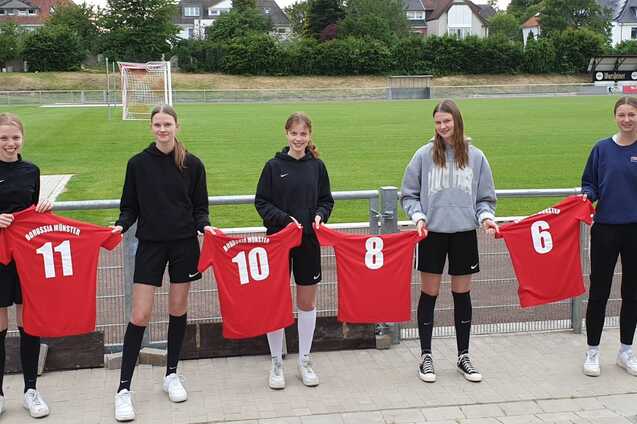 F&uuml;nf Sportlerinnen in Fu&szlig;balltrikots halten rote Shirts mit den Nummern 11, 10, 8 und 6 auf einem Fu&szlig;ballplatz.
