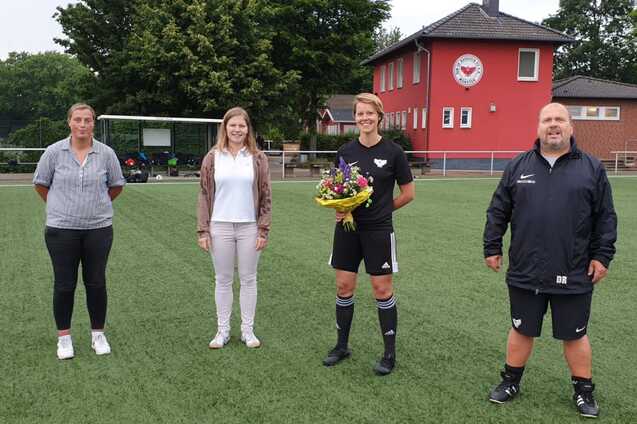Vier Personen stehen auf einem Fu&szlig;ballfeld, eine Spielerin h&auml;lt einen Blumenstrau&szlig;, im Hintergrund ein rotes Vereinsgeb&auml;ude.