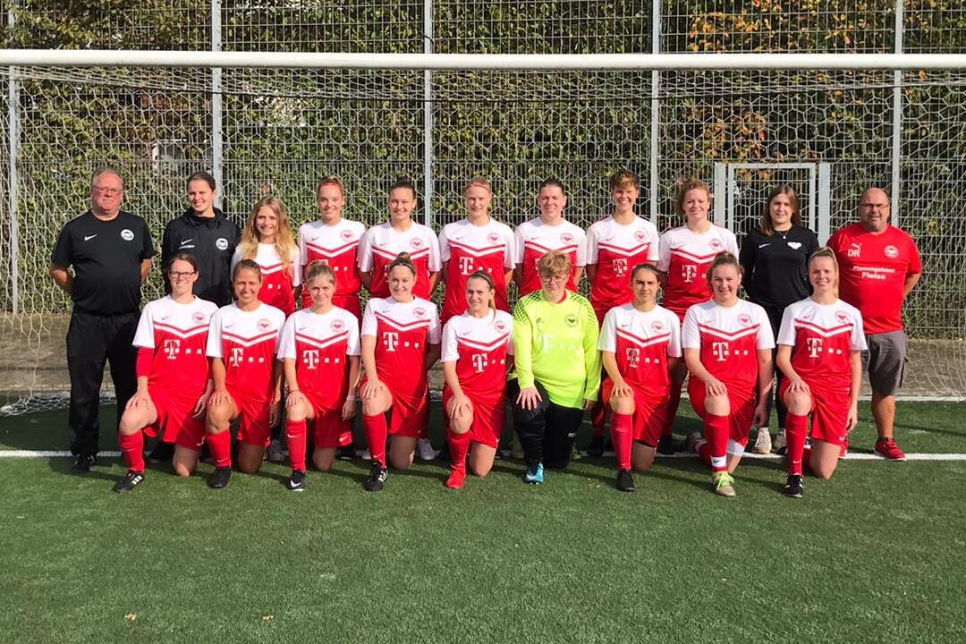 Mannschaftsbild einer Frauenfu&szlig;ballmannschaft in roten Trikots vor einem Tor, begleitet von Trainern auf einem Kunstrasenplatz.