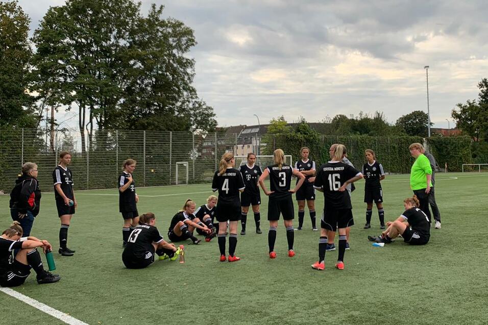 Teambesprechung einer Frauenfu&szlig;ballmannschaft auf einem gr&uuml;nen Kunstrasenplatz unter wolkigem Himmel.
