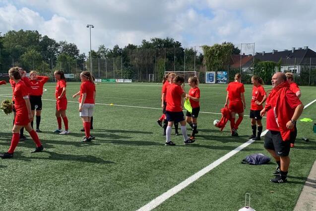 Gruppentraining eines Fu&szlig;ballteams in roten Trikots auf einem Kunstrasenplatz. Spielerinnen stehen und unterhalten sich.