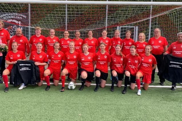 Gruppe von 20 Frauen in roten Fu&szlig;balltrikots pose vor einem Fu&szlig;balltor, einige halten Jacken und Blumen.