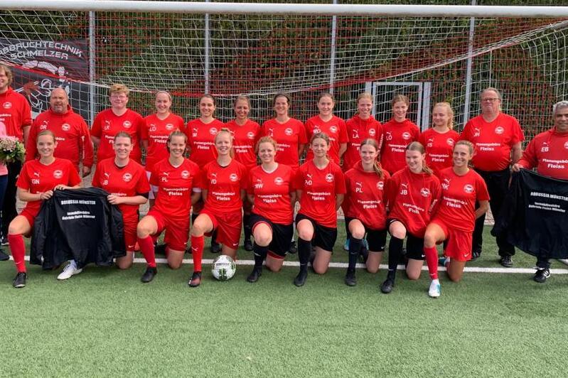Gruppe von 20 Frauen in roten Fu&szlig;balltrikots pose vor einem Fu&szlig;balltor, einige halten Jacken und Blumen.