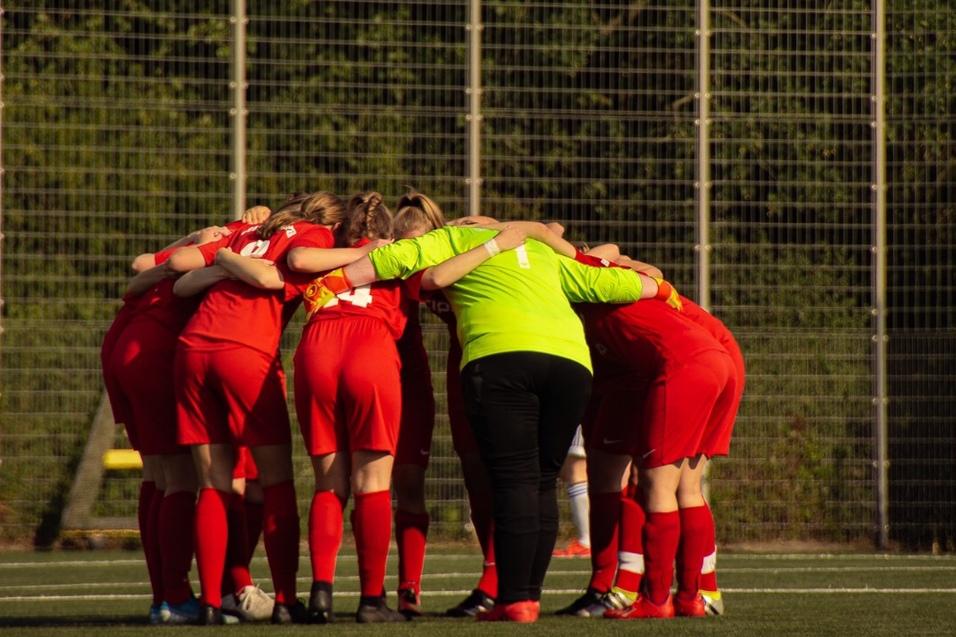 Mannschaft in roten Trikots bildet einen Kreis auf dem Spielfeld, w&auml;hrend ein Schiedsrichter im Hintergrund steht.