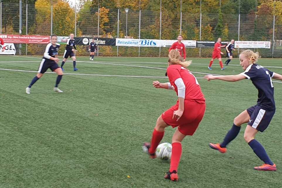 Zwei Fu&szlig;ballspielerinnen in roten und blauen Trikots k&auml;mpfen um den Ball auf einem Kunstrasenplatz.
