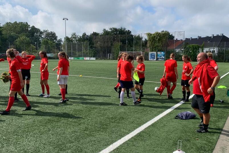 Gruppentraining eines Fu&szlig;ballteams in roten Trikots auf einem Kunstrasenplatz. Spielerinnen stehen und unterhalten sich.