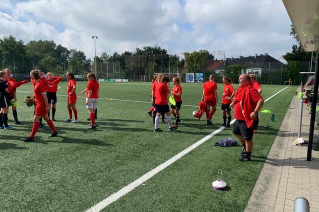 Gruppentraining eines Fu&szlig;ballteams in roten Trikots auf einem Kunstrasenplatz. Spielerinnen stehen und unterhalten sich.