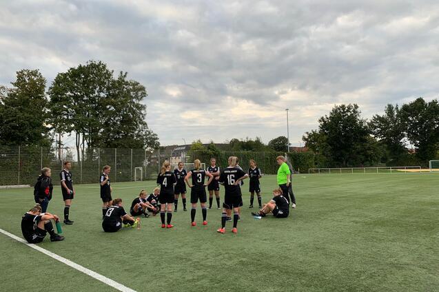 Teambesprechung einer Frauenfu&szlig;ballmannschaft auf einem gr&uuml;nen Kunstrasenplatz unter wolkigem Himmel.