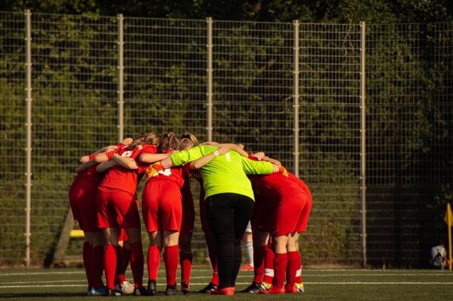 Mannschaft in roten Trikots bildet einen Kreis auf dem Spielfeld, w&auml;hrend ein Schiedsrichter im Hintergrund steht.