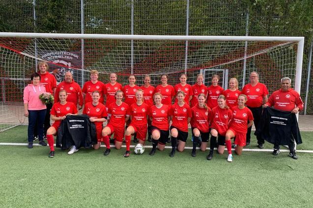 Gruppe von 20 Frauen in roten Fu&szlig;balltrikots pose vor einem Fu&szlig;balltor, einige halten Jacken und Blumen.