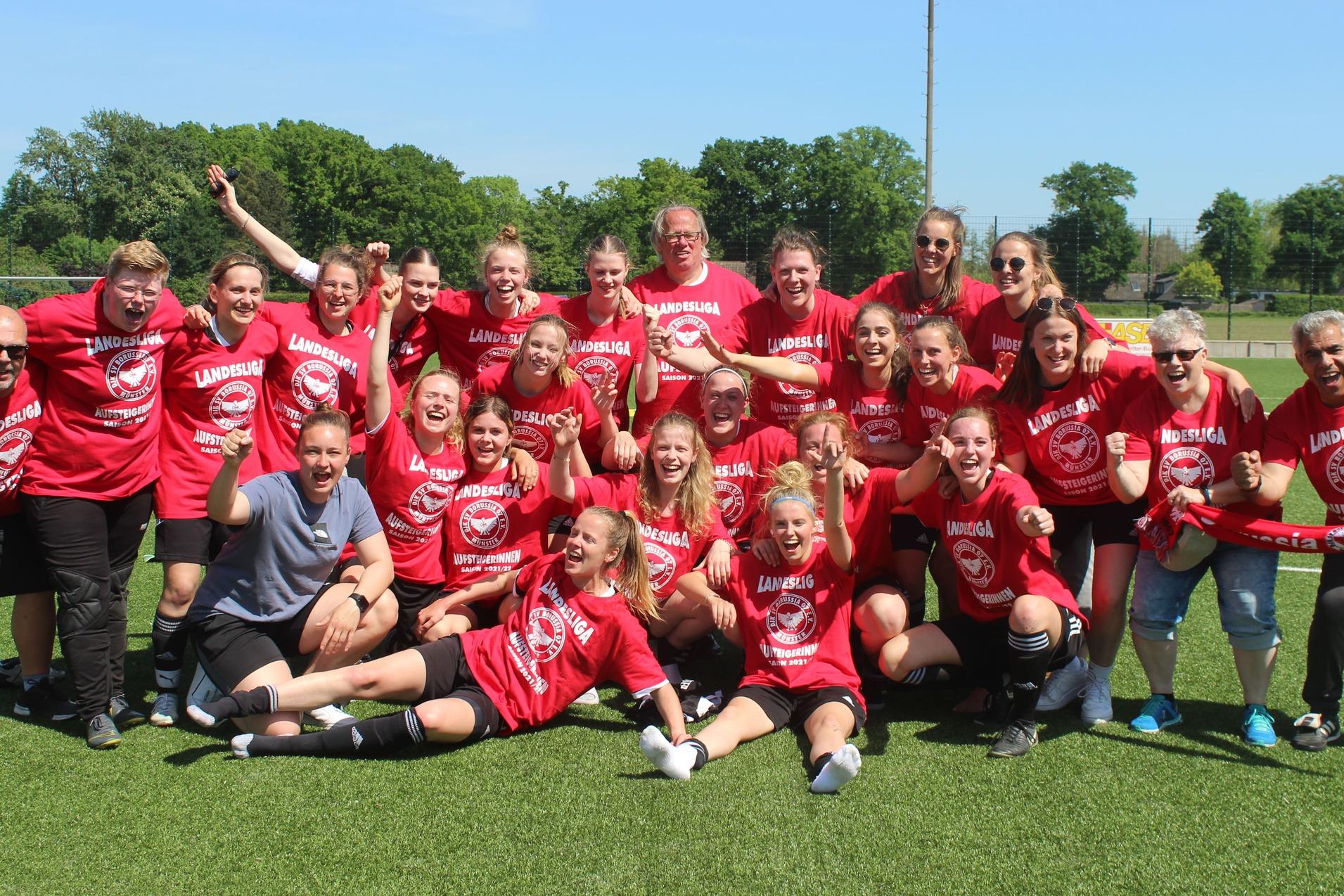 Gruppenshot eines begeisterten Frauenfu&szlig;ballteams in roten Shirts, das auf einem Platz feiert und jubelt.