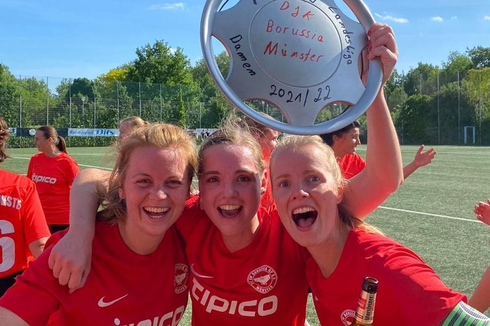 Drei jubelnde Fu&szlig;ballerinnen in roten Trikots halten einen Pokal auf einem Sportplatz und feiern ihren Erfolg.