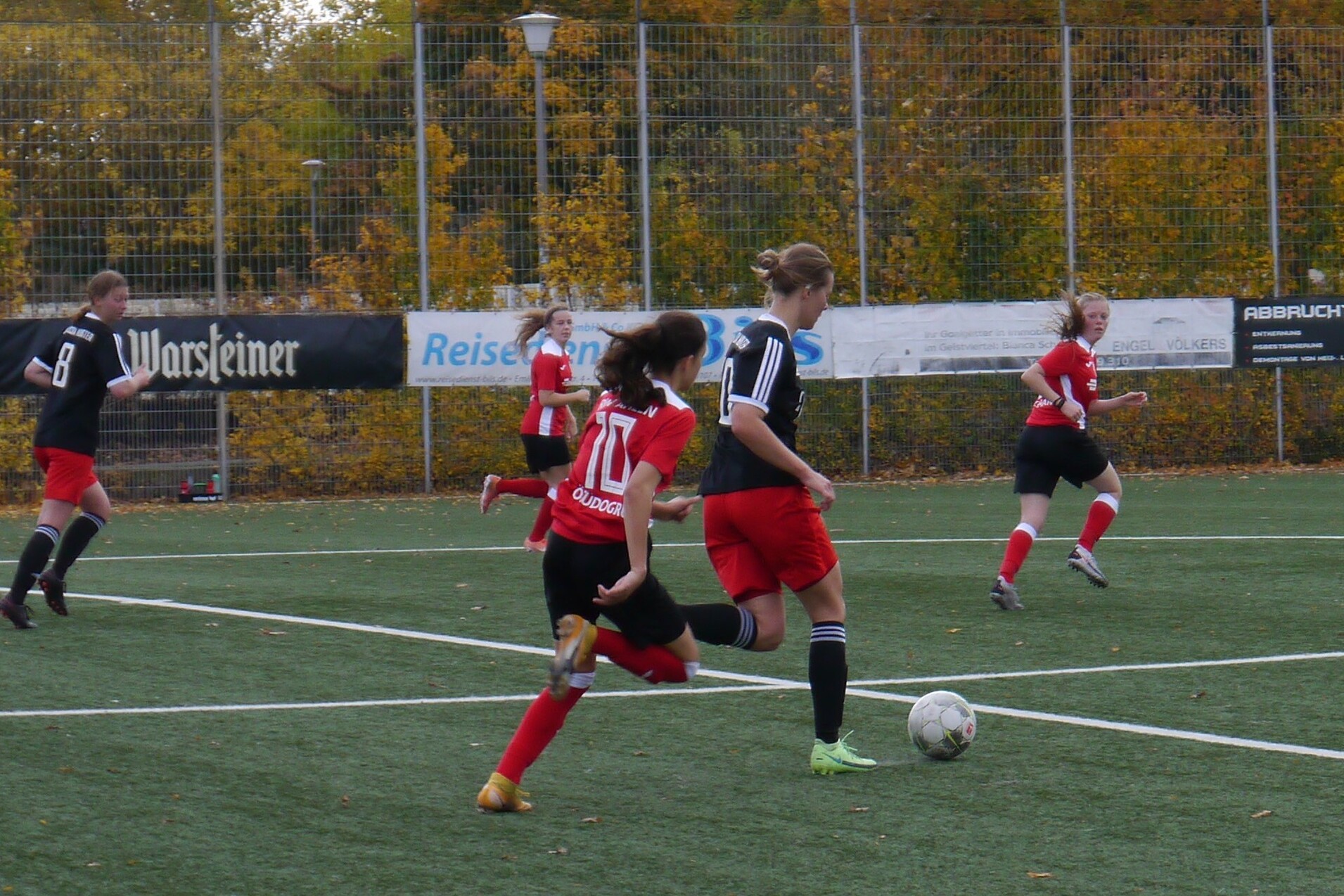 Eine Spielerin mit Ball l&auml;uft auf einem Fu&szlig;ballfeld, w&auml;hrend andere Spielerinnen in roten und schwarzen Trikots zuschauen.