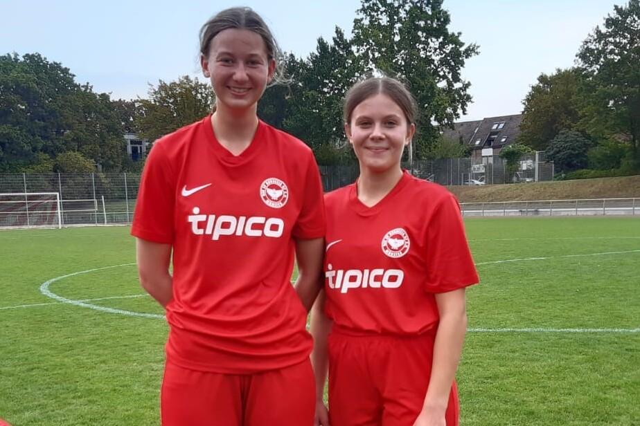 Zwei Fu&szlig;ballspielerinnen in roten Trikots stehen auf einem Rasenplatz, w&auml;hrend eine Dritte sich um die Schuhe k&uuml;mmert.