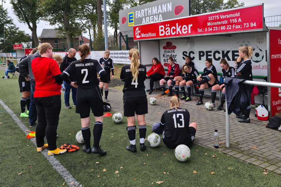 Mannschaftsbesprechung auf einem Fu&szlig;ballplatz, Spielerinnen in Schwarz sitzen auf der Bank, Trainerin spricht mit ihnen.