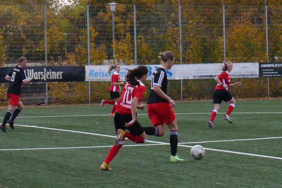 Eine Spielerin mit Ball l&auml;uft auf einem Fu&szlig;ballfeld, w&auml;hrend andere Spielerinnen in roten und schwarzen Trikots zuschauen.