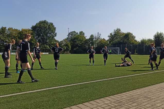 Gruppenspiel auf einem Fu&szlig;ballplatz mit Spielern in schwarzen Trikots, einer liegt auf dem Boden, andere stehen um ihn herum.