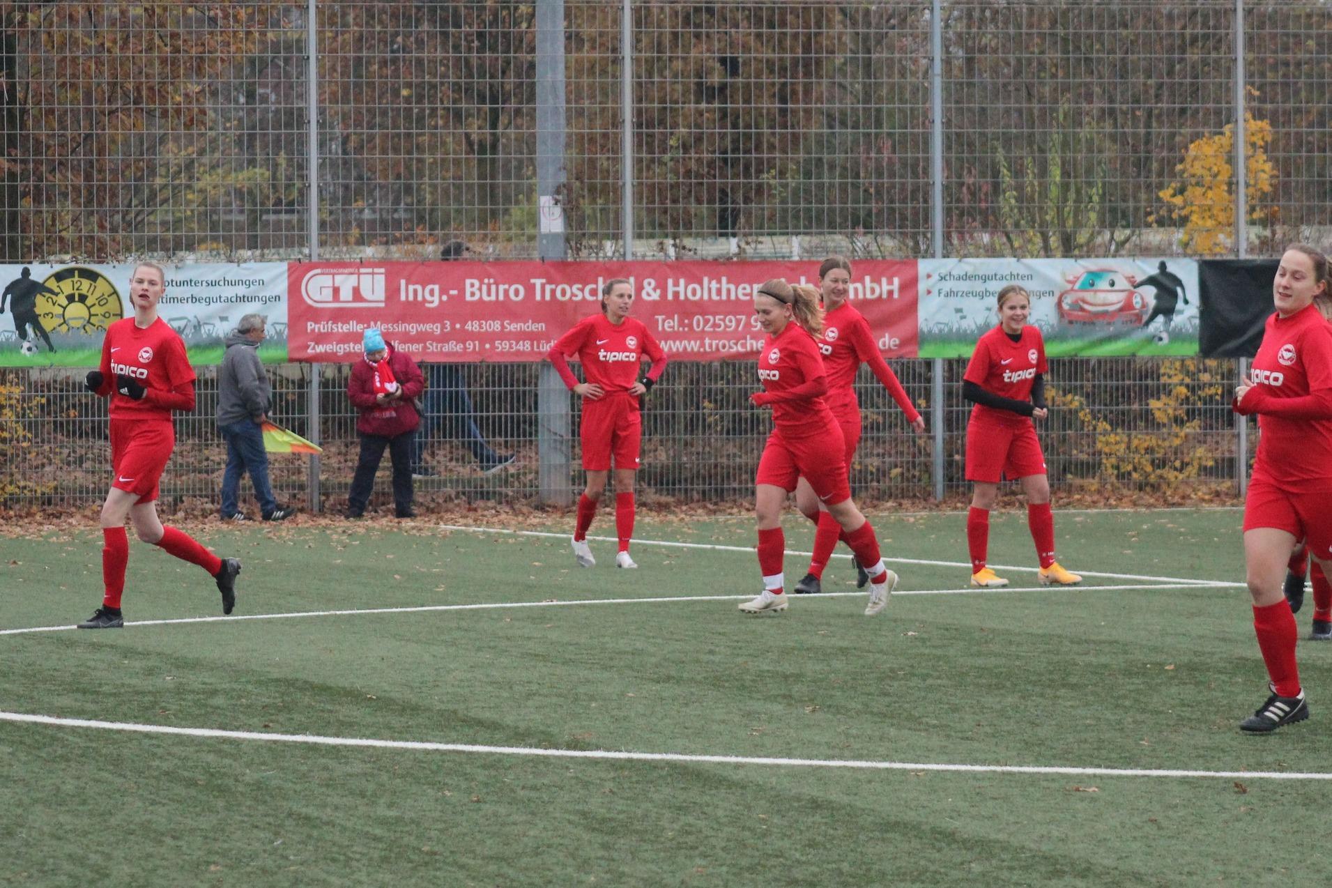 Sechs Fu&szlig;ballspielerinnen in roten Trikots laufen auf einem Kunstrasenplatz, umgeben von Herbstb&auml;umen und Werbebannern.