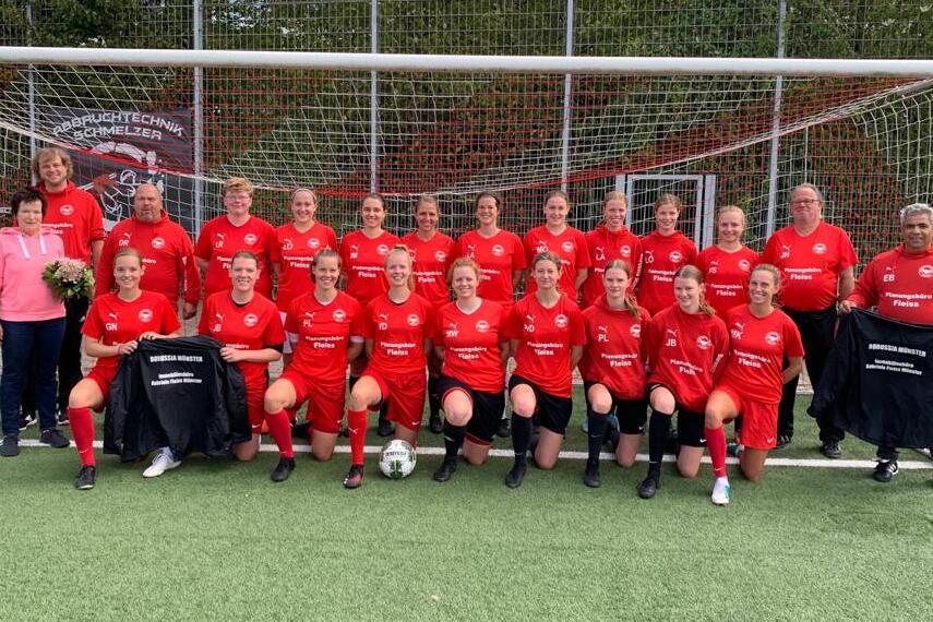 Gruppensfoto der Frauenfu&szlig;ballmannschaft in roten Trikots vor einem Tor auf einem Kunstrasenplatz.