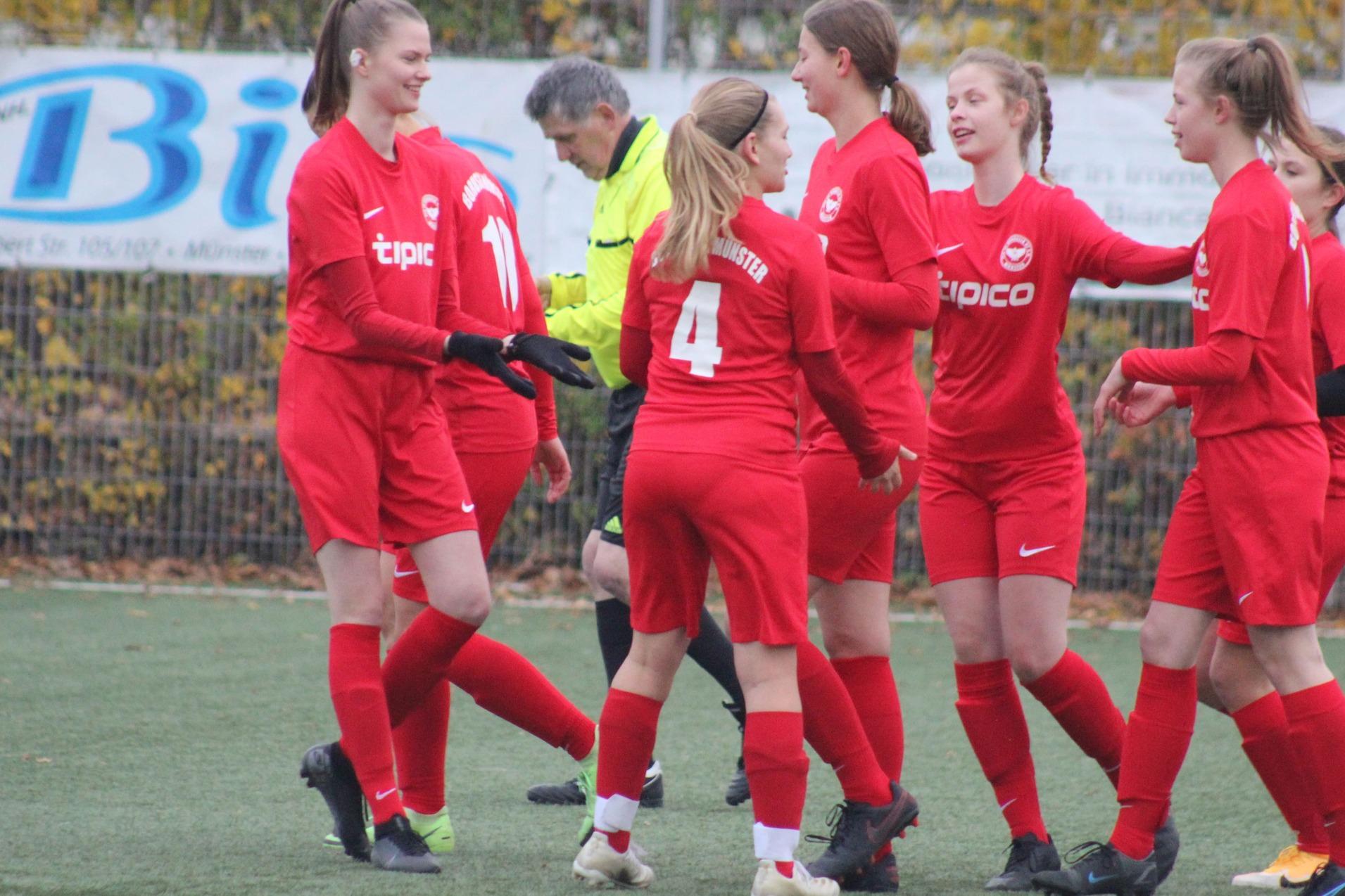 Frauenfu&szlig;ballmannschaft in roten Trikots feiert auf dem Spielfeld, umgeben von gr&uuml;ner Rasenfl&auml;che und einem Schiedsrichter.