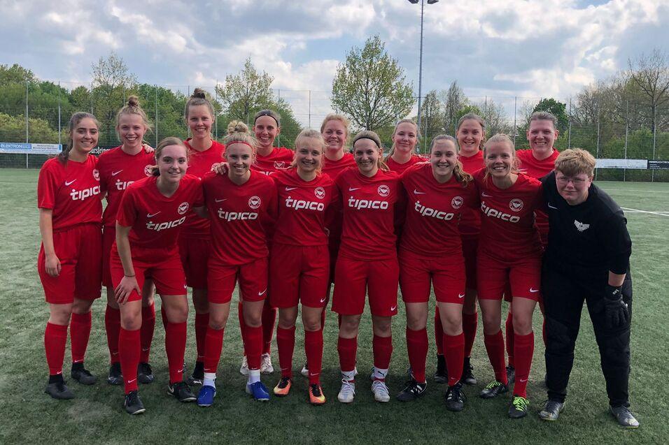 Gruppenfoto einer Frauenfu&szlig;ballmannschaft in roten Trikots auf einem Sportplatz, umgeben von B&auml;umen und einem blauen Himmel.