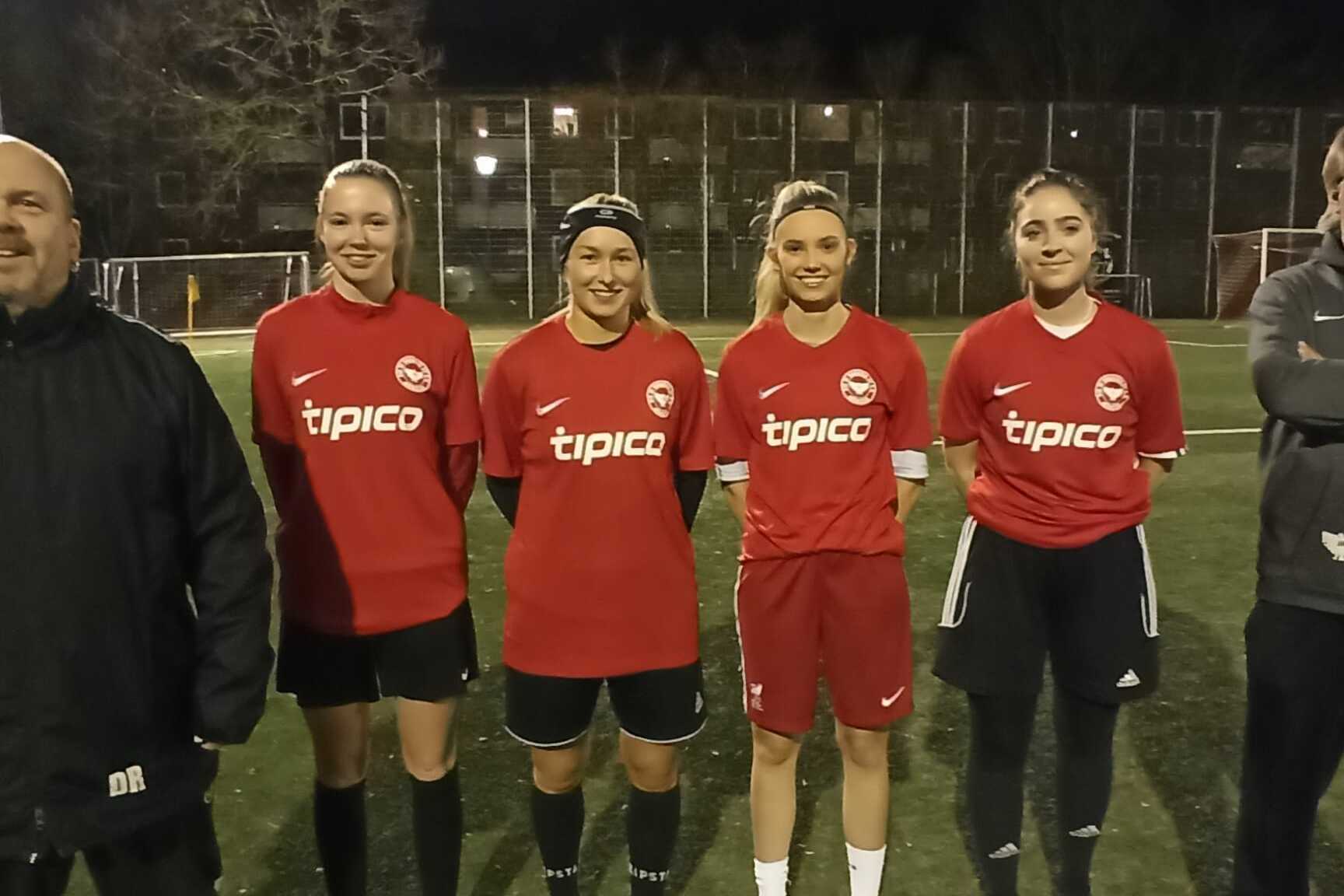F&uuml;nf Fu&szlig;ballspielerinnen in roten Trikots stehen auf einem Sportplatz bei Nacht, zwei Trainer daneben.