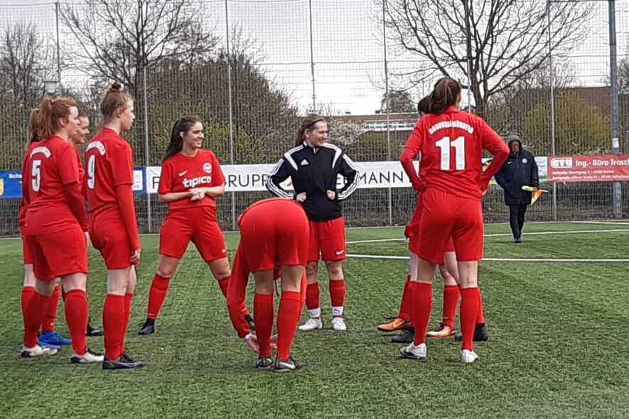 Eine Gruppe von Fu&szlig;ballspielerinnen in roten Trikots versammelt sich auf einem Kunstrasenplatz zu einem Gespr&auml;ch.