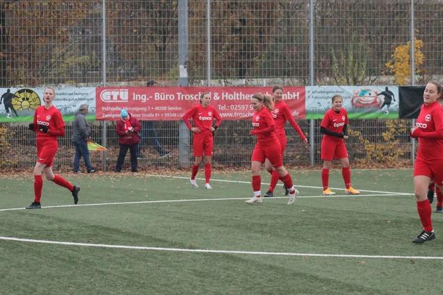Sechs Fu&szlig;ballspielerinnen in roten Trikots laufen auf einem Kunstrasenplatz, umgeben von Herbstb&auml;umen und Werbebannern.