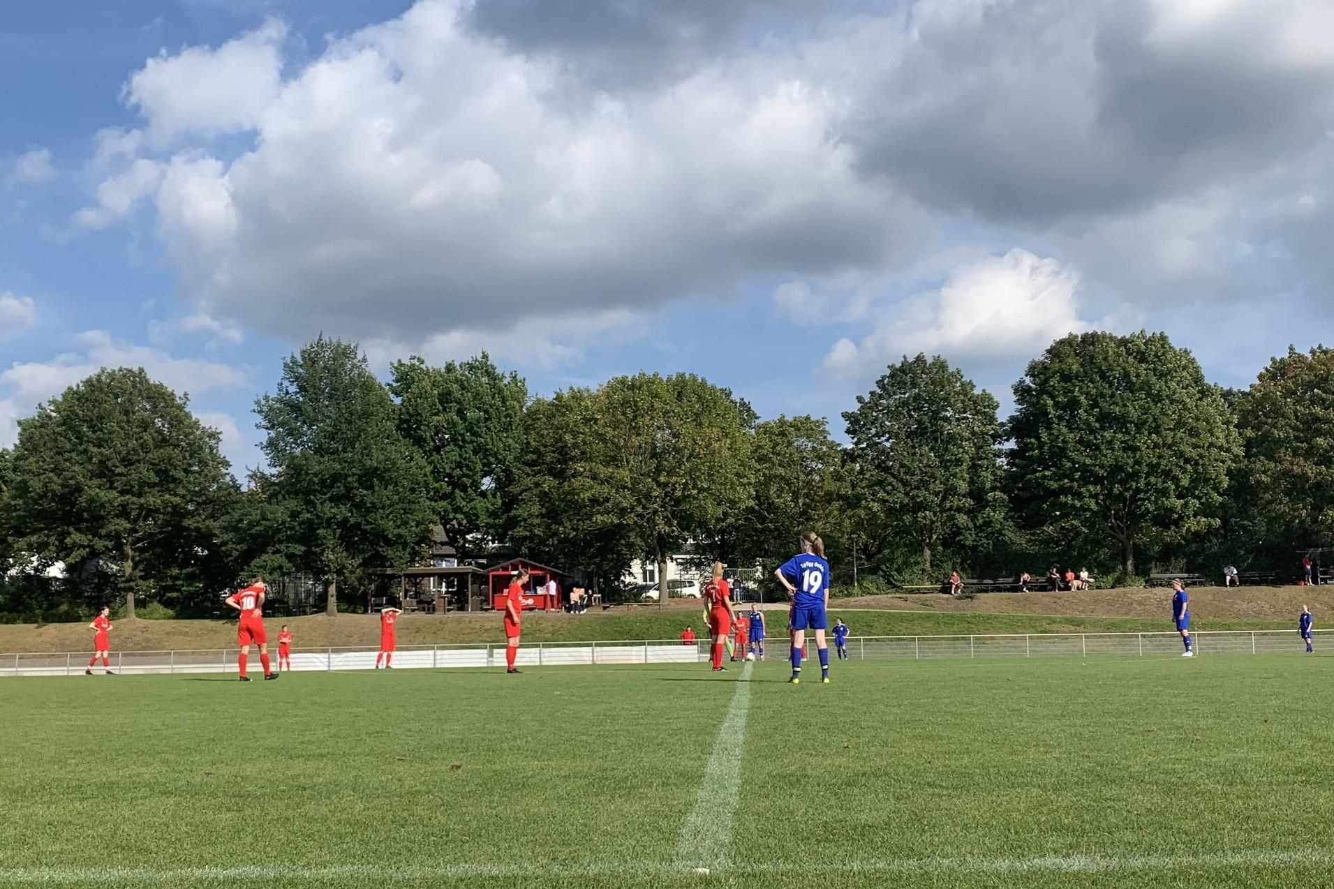 Fu&szlig;ballspiel auf einem Rasenplatz mit Spielern in roten und blauen Trikots unter einem bew&ouml;lkten Himmel.