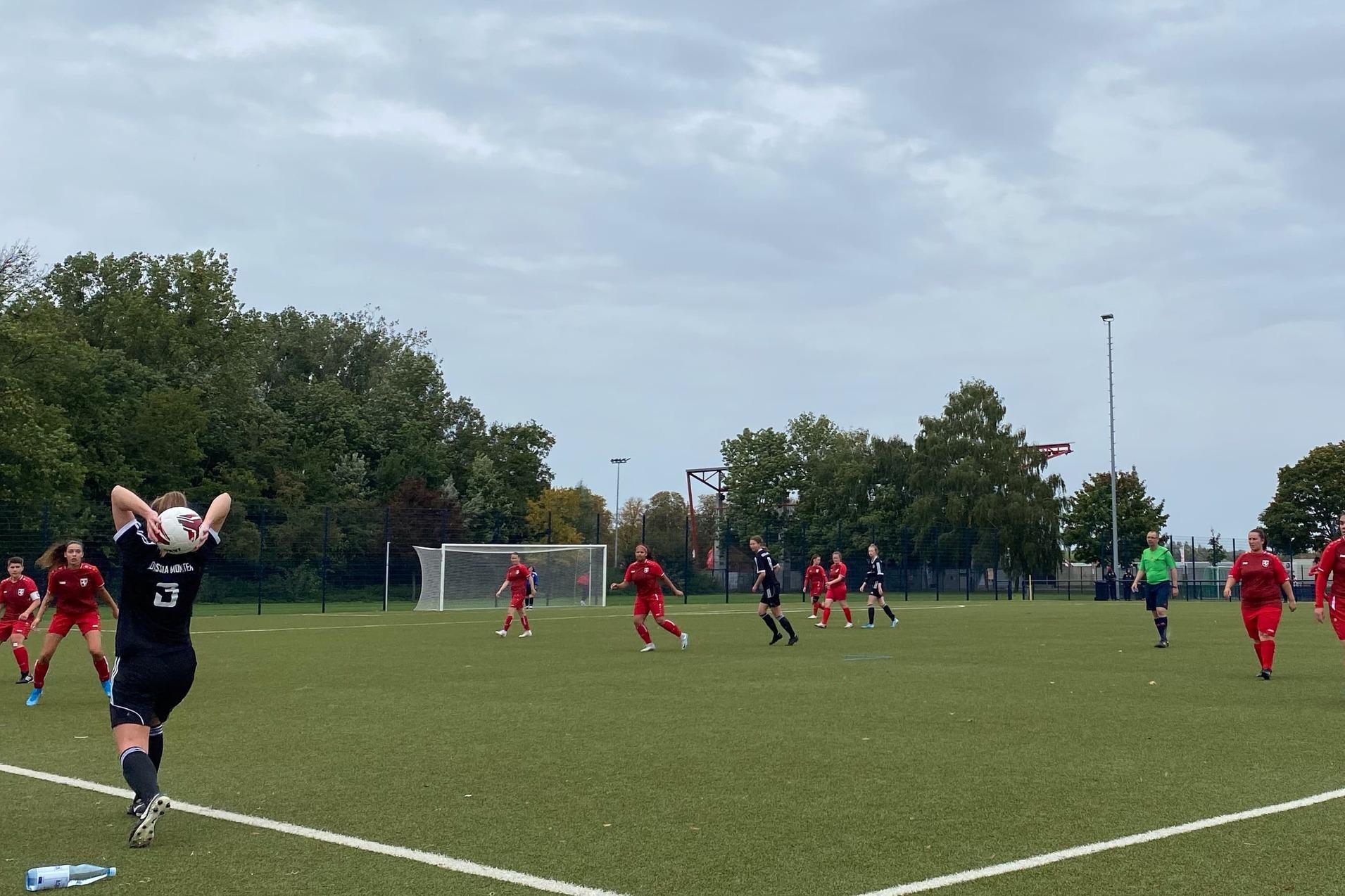 Spielszene auf einem Fu&szlig;ballplatz: Spieler in roten Trikots und ein Gegner mit Ball, im Hintergrund B&auml;ume und &Uuml;berdachung.
