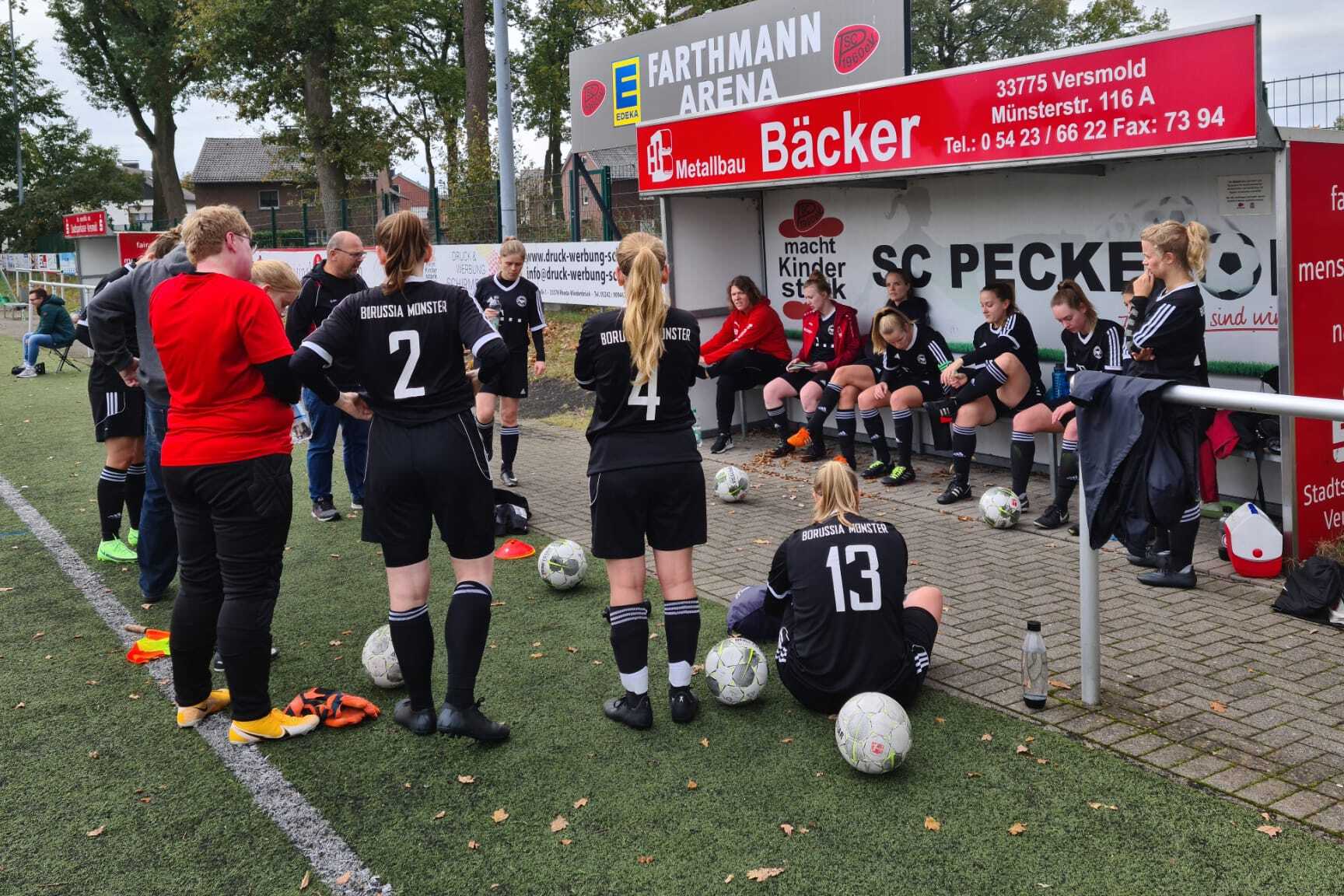 Mannschaftsbesprechung auf einem Fu&szlig;ballplatz, Spielerinnen in Schwarz sitzen auf der Bank, Trainerin spricht mit ihnen.