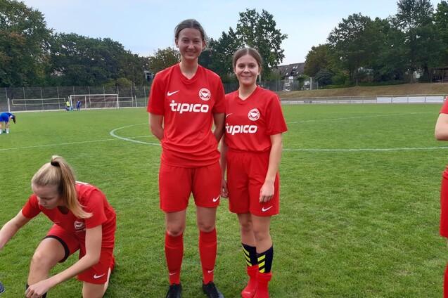 Zwei Fu&szlig;ballspielerinnen in roten Trikots stehen auf einem Rasenplatz, w&auml;hrend eine Dritte sich um die Schuhe k&uuml;mmert.