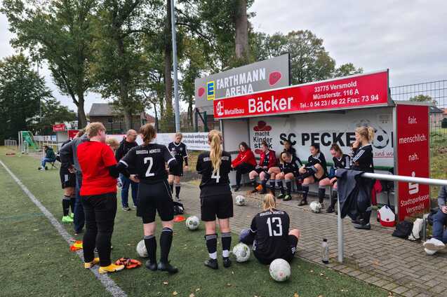 Mannschaftsbesprechung auf einem Fu&szlig;ballplatz, Spielerinnen in Schwarz sitzen auf der Bank, Trainerin spricht mit ihnen.