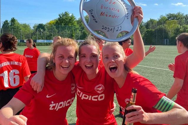 Drei jubelnde Fu&szlig;ballerinnen in roten Trikots halten einen Pokal auf einem Sportplatz und feiern ihren Erfolg.