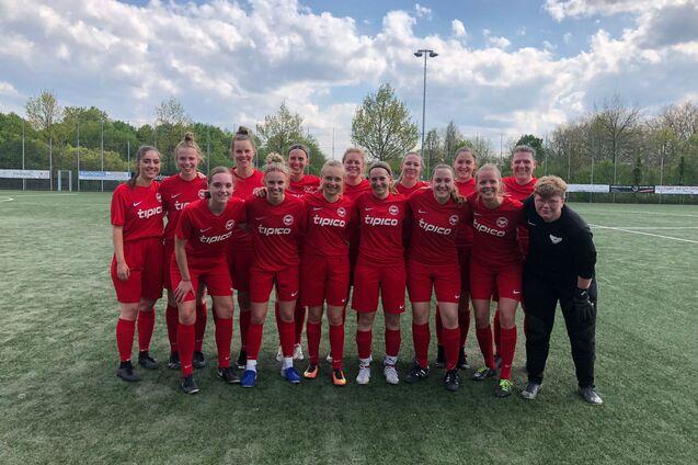 Gruppenfoto einer Frauenfu&szlig;ballmannschaft in roten Trikots auf einem Sportplatz, umgeben von B&auml;umen und einem blauen Himmel.