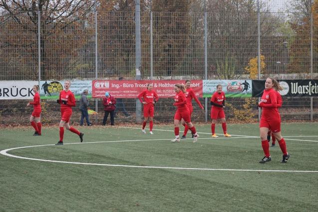 Sechs Fu&szlig;ballspielerinnen in roten Trikots laufen auf einem Kunstrasenplatz, umgeben von Herbstb&auml;umen und Werbebannern.