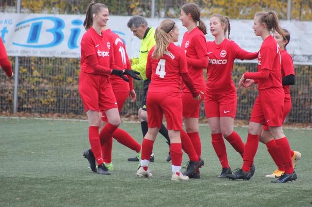 Frauenfu&szlig;ballmannschaft in roten Trikots feiert auf dem Spielfeld, umgeben von gr&uuml;ner Rasenfl&auml;che und einem Schiedsrichter.