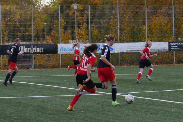 Eine Spielerin mit Ball l&auml;uft auf einem Fu&szlig;ballfeld, w&auml;hrend andere Spielerinnen in roten und schwarzen Trikots zuschauen.