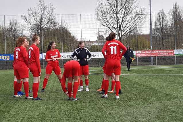 Eine Gruppe von Fu&szlig;ballspielerinnen in roten Trikots versammelt sich auf einem Kunstrasenplatz zu einem Gespr&auml;ch.