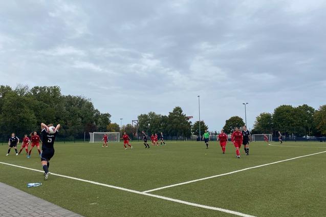 Spielszene auf einem Fu&szlig;ballplatz: Spieler in roten Trikots und ein Gegner mit Ball, im Hintergrund B&auml;ume und &Uuml;berdachung.