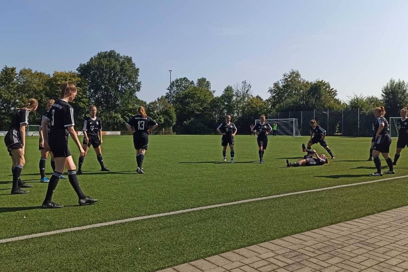Gruppenspiel auf einem Fu&szlig;ballplatz mit Spielern in schwarzen Trikots, einer liegt auf dem Boden, andere stehen um ihn herum.