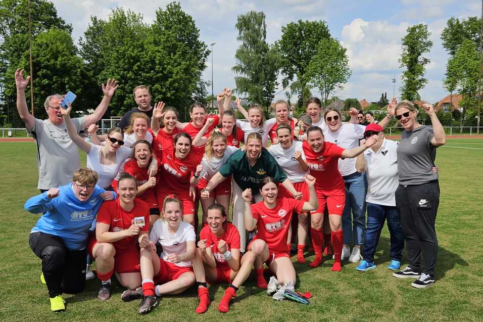 Gruppe von Spielerinnen und Trainern feiert auf einem Fu&szlig;ballplatz, umgeben von B&auml;umen, sonniger Himmel.